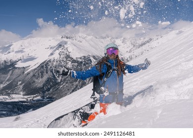 青いジャケット、ヘルメット、マスクを着用した幸せな女性スノーボーダーが、高山の雪崩が発生しやすい地域で雪を投げて楽しんでいます。スキーリゾートでのフリーライド、山頂の景色