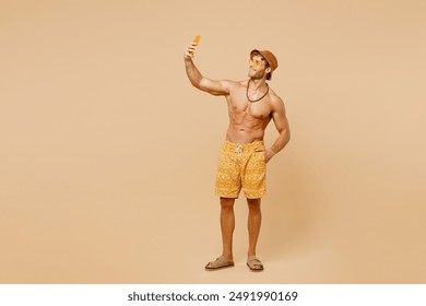 Chàng trai trẻ toàn thân mặc quần short màu vàng đội mũ thư giãn nghỉ ngơi gần hồ bơi khách sạn chụp ảnh tự sướng bằng điện thoại di động đăng ảnh bị cô lập trên nền be đơn giản. Khái niệm tắm nắng biển mùa hè nóng bức