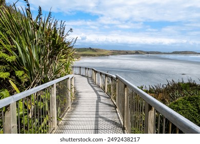 A wooden pathway winds through lush coastal vegetation, leading to a scenic view of a calm bay and distant hills under a blue sky. The pathway invites exploration and wonder.