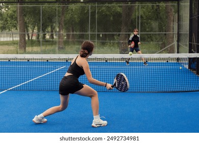 Two young players engaging in a competitive game of padel tennis on a blue outdoor court surrounded by a green park.
