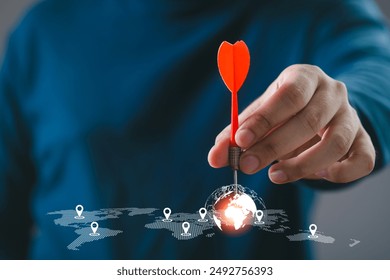 Hand holding a red dart aiming at a glowing world map, symbolizing global target achievement and precision in business strategy.