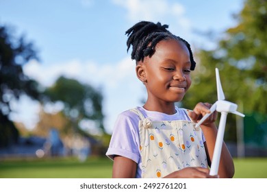Bambina di colore in abiti casual in piedi al parco mentre gioca con le ali di una turbina eolica. Ritratto di una graziosa studentessa che esplora le energie rinnovabili grazie al modellino di una turbina elettrica.