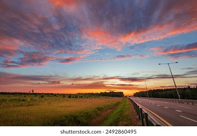 ทางหลวงชนบทที่สวยงามยามพระอาทิตย์ตกดินพร้อมด้วยท้องฟ้าหลากสี เมฆ ทุ่งหญ้า และต้นไม้ที่สร้างฉากอันเงียบสงบ