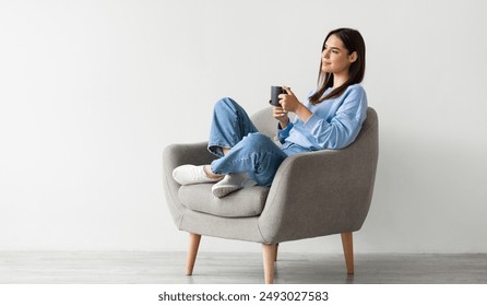 Wanita muda yang positif dalam pakaian kasual duduk di kerusi berlengan dengan kaki bersilang, memanaskan tangan pada secawan kopi panas, menikmati minuman hangat di dinding studio putih, ruang salinan