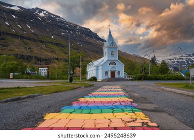 Uma pitoresca igreja azul com uma torre alta em Seyðisfjörður, na Islândia, com um vibrante caminho de paralelepípedos coloridos, nuvens dramáticas e vegetação exuberante.