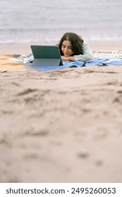 Žena uživa u opuštajućem zimskom danu na plaži, ležeći na ćebetu i gledajući film na svom tabletu