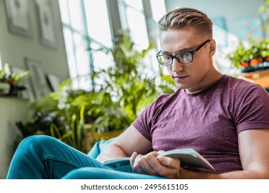 Jovem atraente estudante com óculos focado lendo livro enquanto faz uma pausa no trabalho estudando em um espaço de coworking. Cara descansando relaxando relaxando passando o tempo livre na sala de estar em casa