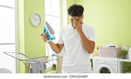 يبدو شاب إسباني محبطًا أثناء حمله مكواة في غرفة الغسيل المضيئة في منزله.