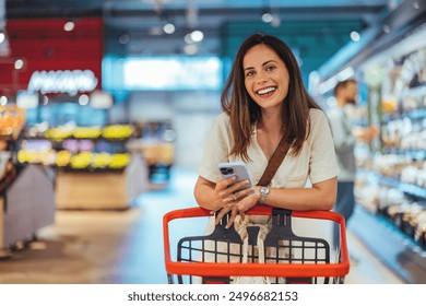 Portrét šťastné ženy nákupu potravin v supermarketu a při pohledu na fotoaparát. Nákup zboží pomocí smartphonu v obchodě s potravinami. Zákaznice nakupování s kontrolním seznamem pro smartphone