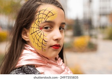 美しい茶色の目をした長い髪の 10 代の女の子が、コスチューム パーティー用に黒いクモとクモの巣が付いた緑の帽子をかぶって微笑みながら下を向いており、頬には水化粧のクモの巣があり、秋の背景はぼやけています。