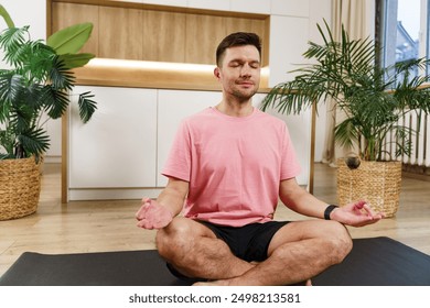 Man in pink shirt meditating on a yoga mat in a modern, plant-filled room with eyes closed.