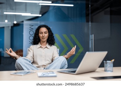 Businesswoman practicing mindfulness meditation in modern office. Woman meditating in lotus position in front of laptop, promoting calm and focus in workplace