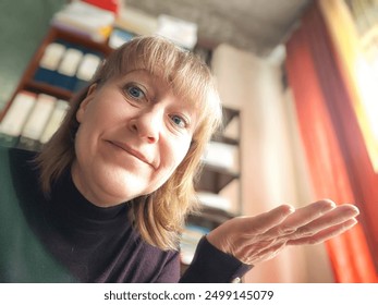 Ženska se nasmehne in iztegne roko na fotografiji od blizu. Ima blond lase, modre oči in nosi vijoličen pulover