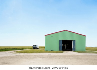 Een groen pakhuis met een rood dak staat in een landelijke omgeving, met een vrachtwagen geparkeerd op een onverharde weg onder een helderblauwe lucht.