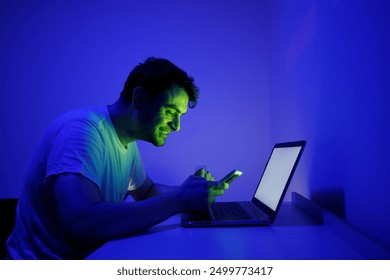 Un giovane in una stanza illuminata di blu davanti al suo computer portatile. Luce verde sul suo viso. Guarda il cellulare e rimane scioccato.