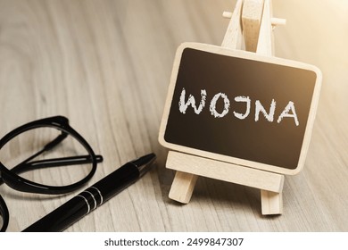 Black writing board on a wooden frame with the inscription "Wojna", next to black glasses and a pen (selective focus) translation: WAR