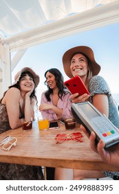 Vertical young group of only women paying the bill with a nfc cellphone in cocktail beach bar. Female holding a smart phone and giving a payment transaction to the cashier on vacations with friends