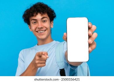 Confident boy guy high school male pupil student teenager with dental braces backpack holding showing smart mobile phone`s screen mock up pointing index finger on you isolated over blue background