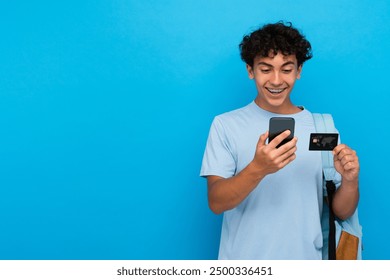 Garoto feliz e animado, aluno do ensino médio, estudante adolescente com mochila segurando cartão de crédito bancário de plástico e telefone celular inteligente usando aplicativo bancário on-line isolado no espaço de cópia de fundo azul