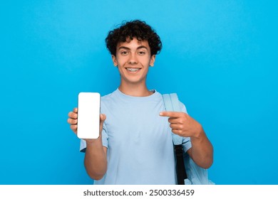 Happy smiling boy guy high school male pupil student teenager with backpack pointing index finger at smart mobile cell phone showing mock up screen isolated on blue background. Education concept