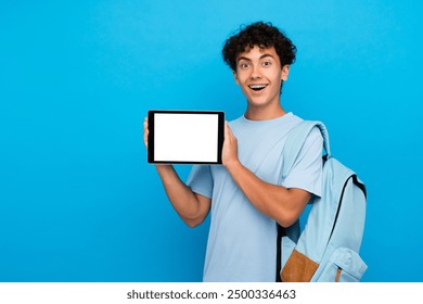 Garoto feliz surpreso, aluno do ensino médio, estudante adolescente com bolsa segurando um tablet digital para e-learning, mostrando, demonstrando, simulando, tela branca vazia, isolada sobre fundo azul