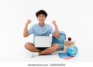 Garoto feliz vencedor, aluno do ensino médio, estudante adolescente sentado perto de livros, cadernos, mochila, globo, usando laptop, gesto de vitória, comemorando o sucesso, isolado no fundo branco.