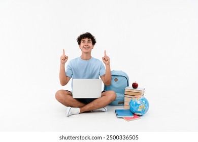 Garoto feliz, aluno do ensino médio, estudante adolescente sentado perto de livros, cadernos, mochila, globo, segurando, usando, laptop, para, e-learning, apontando, dedo indicador, isolado, branca, fundo.