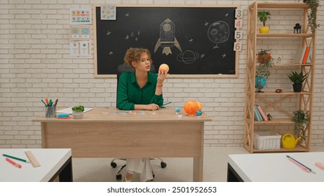 Lelaki dan perempuan sekolah rendah duduk di dalam bilik dalam kelas astronomi. Guru wanita memegang model planet dan menerangkannya kepada kanak-kanak. Konsep pendidikan moden.