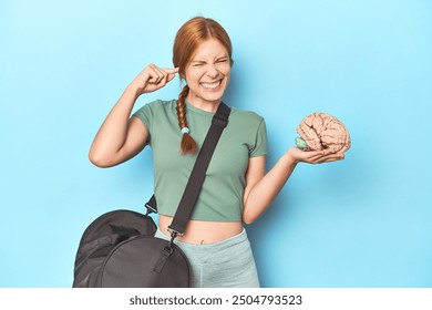 Sporty redhead with brain model on blue background covering ears with hands.