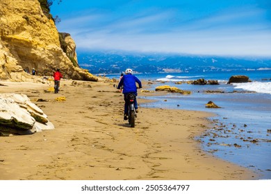 seorang lelaki memakai jaket merah menunggang basikal elektrik di pantai berpasir coklat dengan orang ramai berjalan di sepanjang pantai, air laut biru dan pokok hijau yang subur di Pantai Leadbetter di Santa Barbara California
