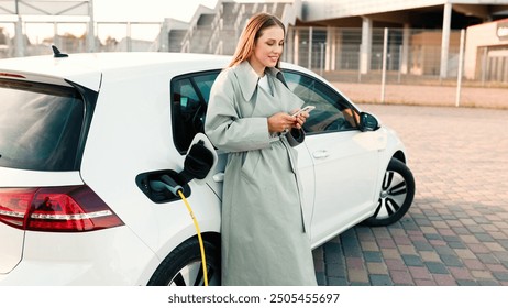 Un prim plan al mașinii electrice albe se încarcă în aer liber în oraș într-o zi însorită, în timp ce femeia atinge smartphone-ul. Vehicul electric la stația de încărcare afară, mâinile femeii folosind telefonul mobil. Conceptul EV