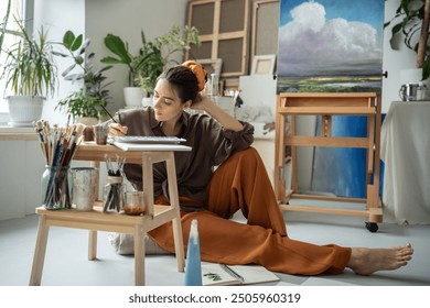 Una artista reflexiva experimenta una crisis de inspiración, reflexiona sobre el dibujo de un álbum y se sienta en el suelo en su estudio de arte, pensando en ideas. Una pintora pensativa en un taller creativo dibujando una ilustración
