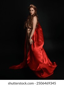 Full length portrait of beautiful female model wearing grecian red silk gold roman robes, historical fantasy goddess costume with crown wreath. Standing pose isolated on dark studio background.