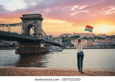 Ciesząc się wakacjami w Budapeszcie. Młoda podróżująca kobieta z węgierską flagą narodową spacerująca po promenadzie nad rzeką z widokiem na miasto o zachodzie słońca