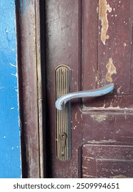 Detailný obrázok starých zvetraných drevených dverí s odlupujúcim sa náterom, s vintage mosadznou kľučkou a zámkom. Kontrastné modré lemovanie dodáva rustikálnemu prostrediu charakter.