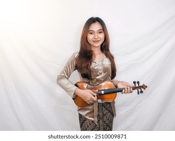 Una bella mujer indonesia con una kebaya tradicional toca alegremente el violín. Mujer asiática con ropa tradicional de Indonesia. Concepto de arte musical