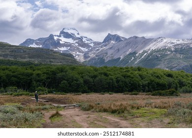 Wanderweg im offenen Feld in Richtung eines Waldes mit vielen Bäumen; einige Leute gehen auf Holzplattformen auf dem Weg; schneebedeckte Berge und bedeckter Himmel