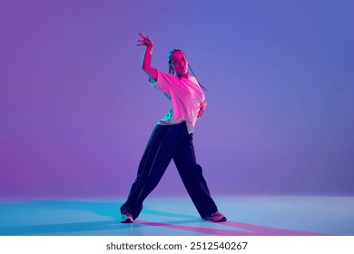 Young beautiful girl in stylish clothes performing hip hop with passion and energy against gradient purple background in neon light. Concept of freestyle dance, modernity, urban culture, youth