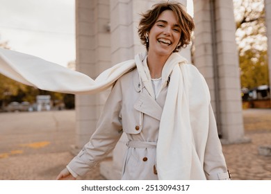 pretty smiling woman walking in park in winter clothes, portrait in beige trench and white knitted scarf, winter cold season accessories trend, stylish outfit, elegant beautiful lady, short haircut