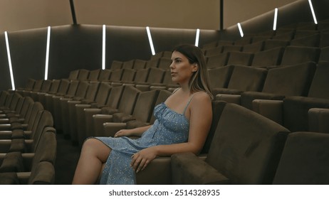 Tânără brunetă în rochie albastră stând singură într-un teatru modern întunecat, arătând gânditoare.