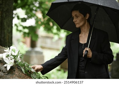 Medium shot of senior woman in mourning veil placing white lilies on stone of memorial monument at grave grieving over loss of loved one, while hiding from rain under black umbrella at cemetery