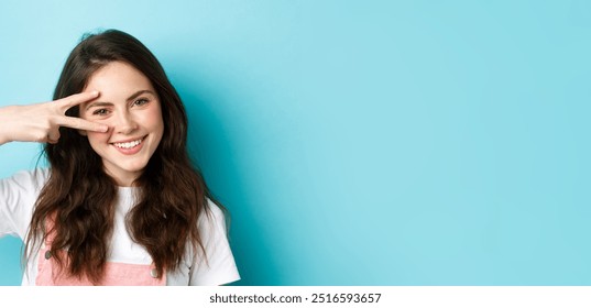 Beauty. Close up of attractive young woman with clean skin and cute blush, showing v-sign near face, smiling with teeth, standing over blue background.