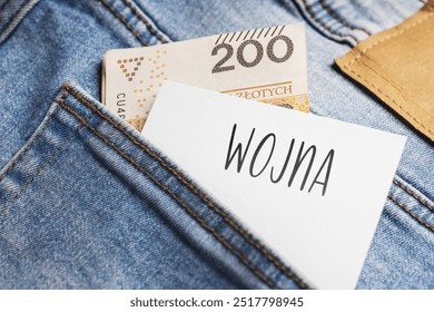 White card with a handwritten inscription "Wojna", put into the pocket of blue pants jeasnow, next to Polish banknotes PLN (selective focus), translation: War