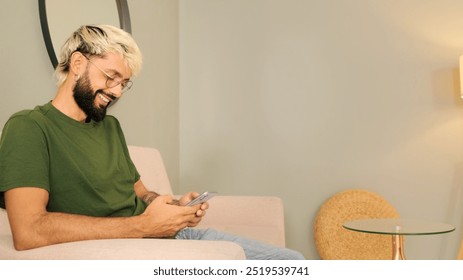 Gros plan, un jeune homme blond avec une barbe portant un t-shirt vert olive se repose assis sur un canapé dans un salon confortable en utilisant un téléphone portable