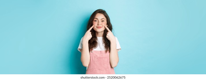 Portrait of beautiful smiling brunette woman with cute blush, pointing at cheeks and looking happy at camera, advertising make up or cosmetics, standing against blue background.
