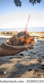 Un fel de mâncare vibrant de „rujak”, o salată de fructe tradițională indoneziană cu sos de arahide picant, este servit pe o plajă cu nisip. Fructele tropicale proaspete contrastează frumos cu marea turcoaz și cu cerul senin