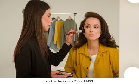 Caucasian woman makeup artist powdering girl trendy blogger in studio reflection female stylist applying shadows visage on influencer face brush getting ready for filming blog cosmetics fashion beauty