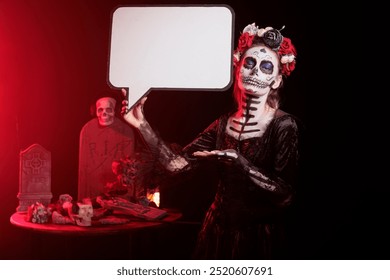 Femeie care acționează ca zeiță a morții ținând un balon de vorbire, purtând machiaj cu craniu pentru evenimentul dios de los muertes. Femeie folosind șablon copyspace izolat pe carton, reclamă santa muerte
