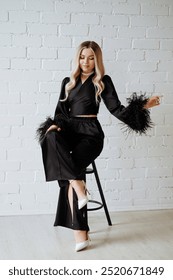 Elegant blonde woman with long hair in black dress with feathers on sleeves sits on high stool against white brick wall
