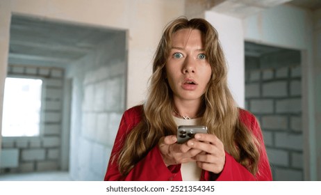 Шокирана жена чете лоши новини на смартфон в недовършен апартамент.